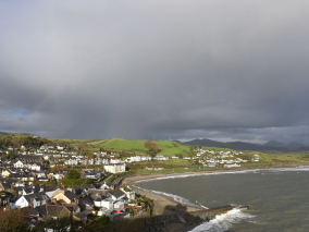 Vista panorâmica de uma cidade costeira com casas agrupadas próximas ao mar, rodeada por colinas verdes e sob um céu nublado com um arco-íris ao fundo